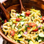 A wooden bowl filled with rotini pasta salad, featuring cherry tomatoes, cucumber, chickpeas, red onion, and herbs, with wooden serving utensils resting inside the bowl.