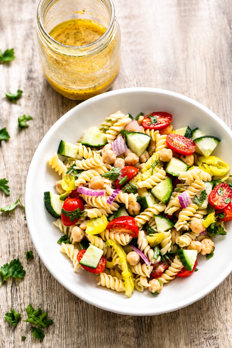 A white bowl filled with pasta salad featuring rotini, cherry tomatoes, cucumber, chickpeas, red onion, and pepperoncini sits on a wooden surface next to a jar of yellow vinaigrette dressing.