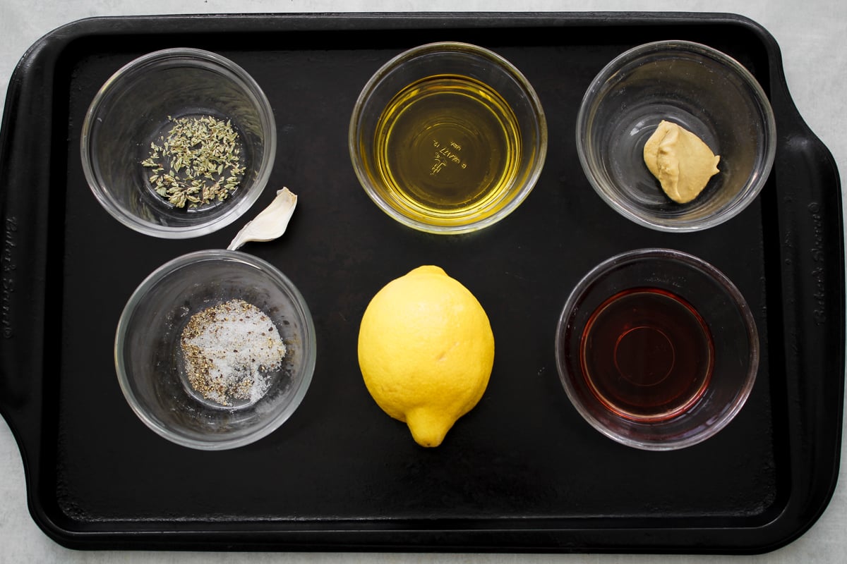 Six small bowls and a lemon on a black tray. The bowls contain dried herbs, olive oil, Dijon mustard, salt and pepper, a garlic clove, and red vinegar, arranged around the whole lemon in the center.