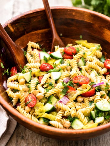 A wooden bowl filled with rotini pasta salad, featuring cherry tomatoes, cucumber, chickpeas, red onion, and herbs, with wooden serving utensils resting inside the bowl.