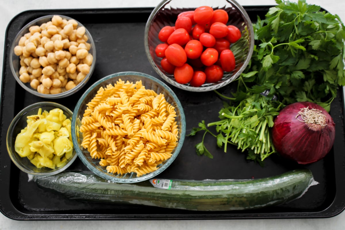 A tray with bowls of chickpeas, grape tomatoes, fusilli pasta, and pepperoncini, alongside fresh parsley, cilantro, a red onion, and a whole cucumber.