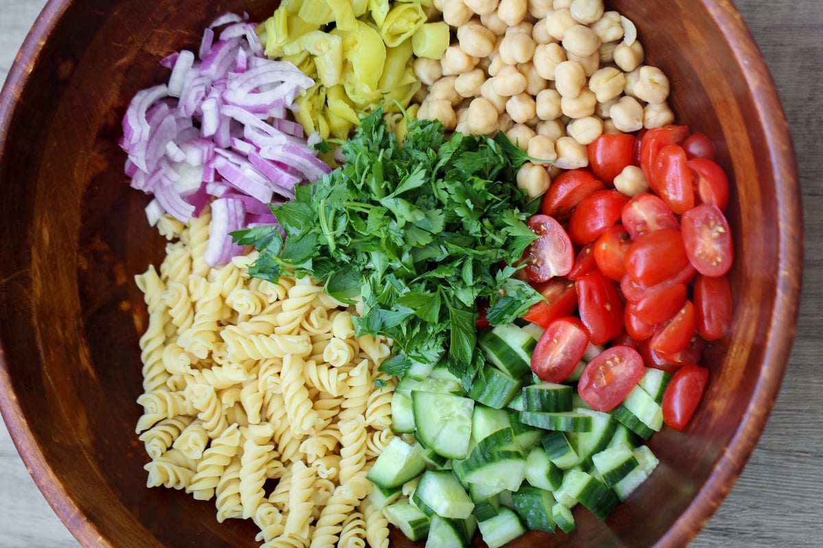 A wooden bowl filled with chopped red onion, artichoke hearts, chickpeas, cherry tomatoes, chopped parsley, diced cucumber, and rotini pasta, arranged in separate sections.