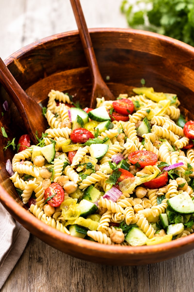A wooden bowl filled with rotini pasta salad, featuring cherry tomatoes, cucumber, chickpeas, red onion, and herbs, with wooden serving utensils resting inside the bowl.