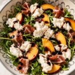 A bowl of arugula salad topped with peach slices, prosciutto, dollops of burrata cheese, and drizzled with balsamic glaze. The salad is arranged in a patterned bowl on a marble surface.