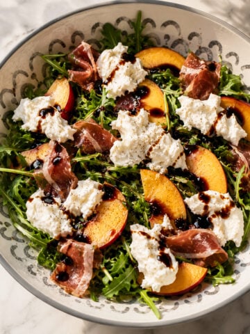 A bowl of arugula salad topped with peach slices, prosciutto, dollops of burrata cheese, and drizzled with balsamic glaze. The salad is arranged in a patterned bowl on a marble surface.