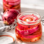 A glass jar filled with pickled red onion slices in brine, with another jar and a whole red onion in the background, all placed on a white marble surface.