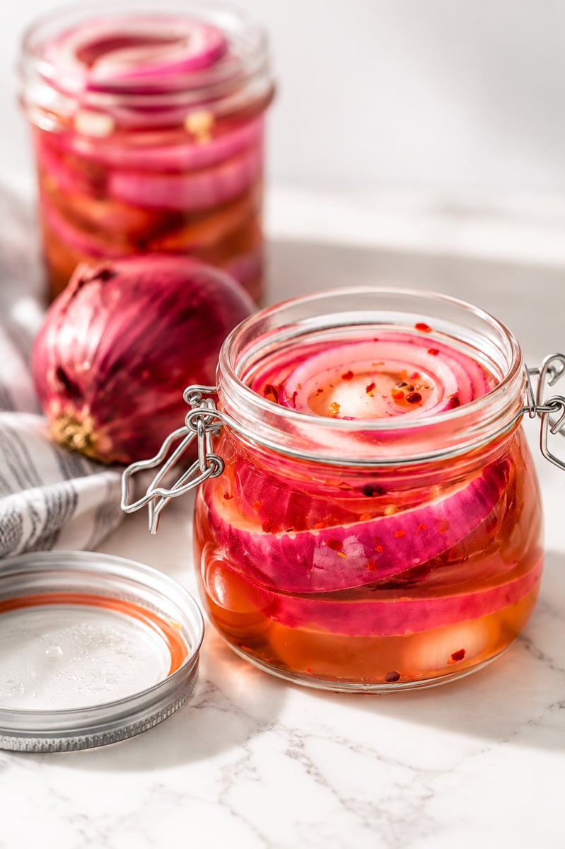 A glass jar filled with pickled red onion slices in brine, with another jar and a whole red onion in the background, all placed on a white marble surface.