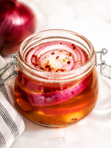 A glass jar filled with pickled red onions in brine, topped with chili flakes, sits on a marble surface next to a red onion and a striped kitchen towel.