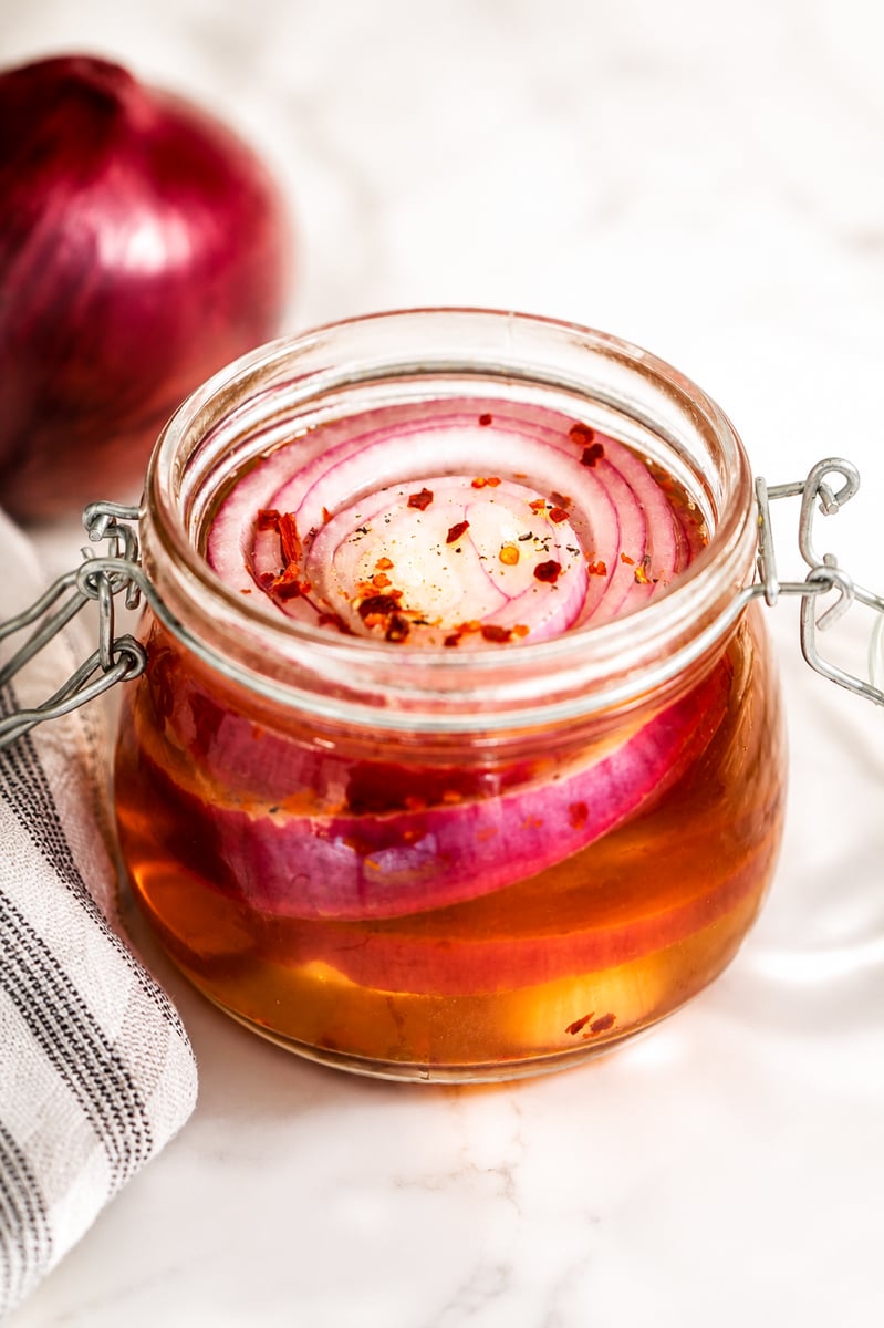 A glass jar filled with pickled red onions in brine, topped with chili flakes, sits on a marble surface next to a red onion and a striped kitchen towel.