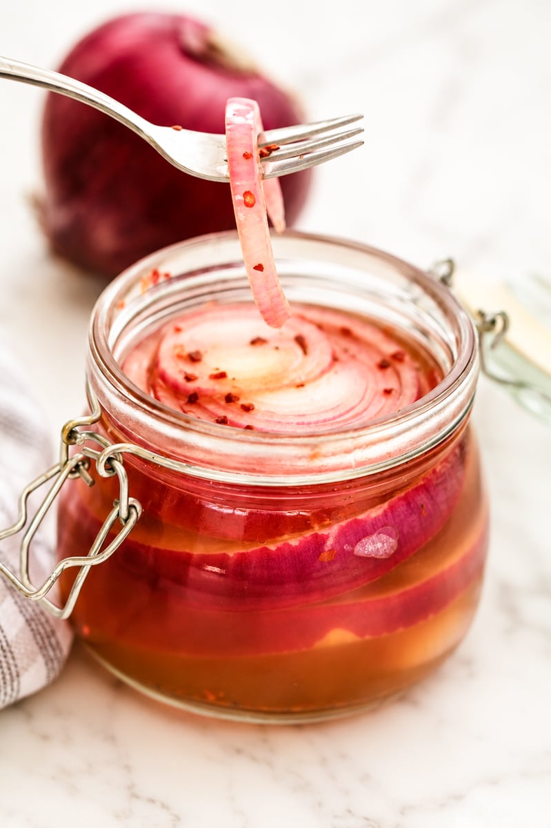A fork lifts a slice of pickled red onion from an open glass jar filled with pickled onions and brine, with a whole red onion in the background on a marble surface.
