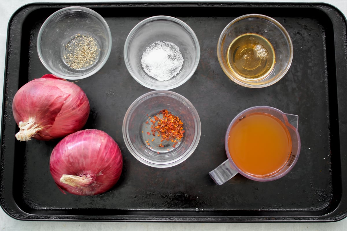 A black tray holds two red onions, a measuring cup of brown liquid, and four small bowls containing salt, black pepper, chili flakes, and a golden liquid.