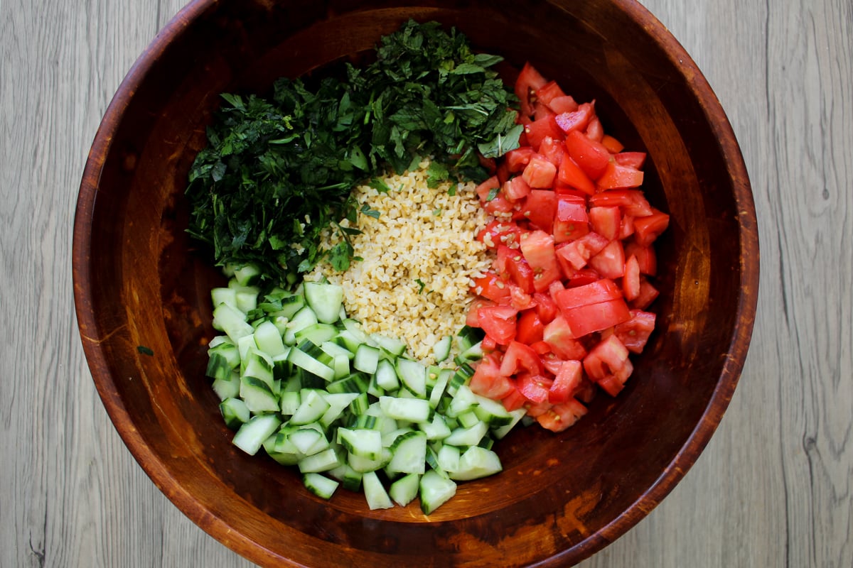 A wooden bowl contains chopped parsley, diced tomatoes, diced cucumbers, and cooked bulgur, arranged in separate sections on a light wood surface.