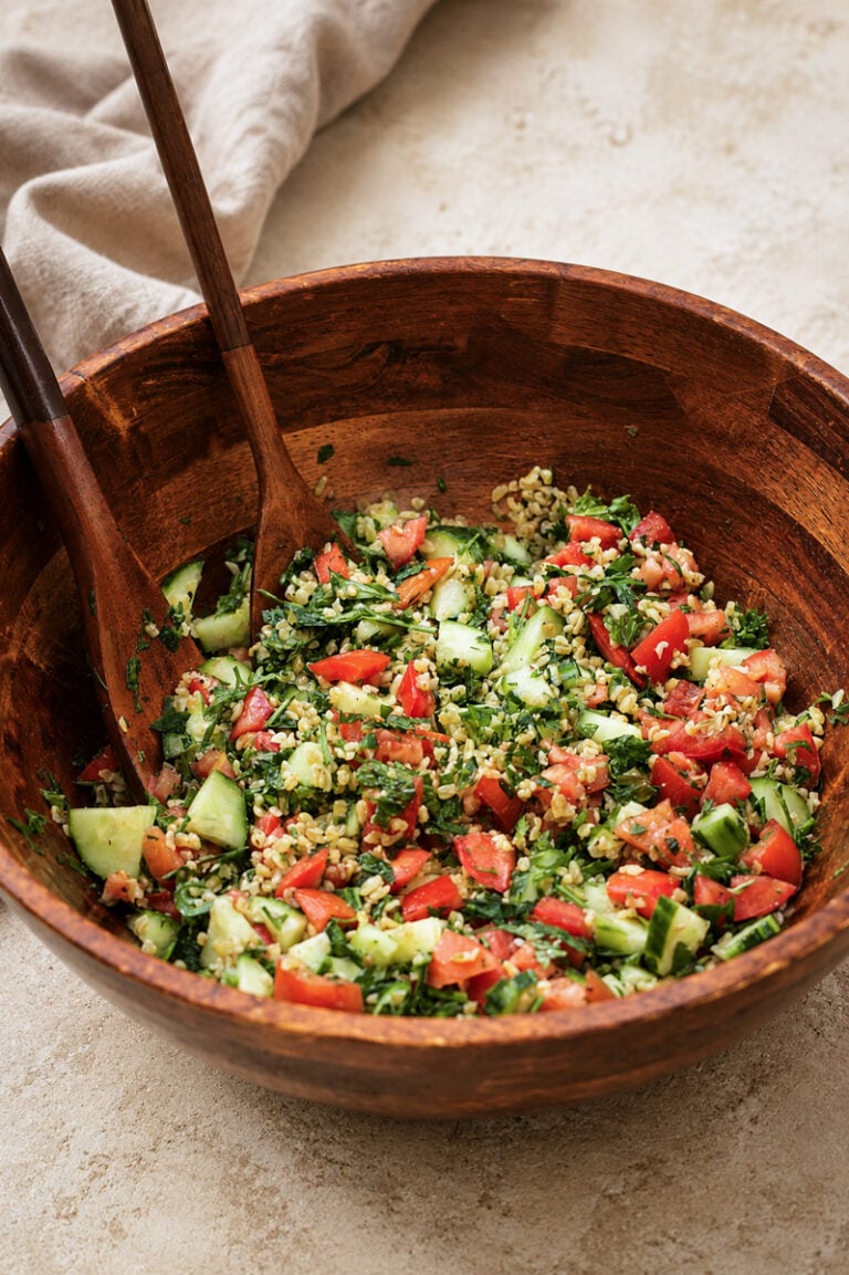 A wooden bowl filled with tabbouleh salad, featuring chopped tomatoes, cucumbers, parsley, and bulgur, with two wooden serving utensils resting inside the bowl. A beige cloth lies in the background.