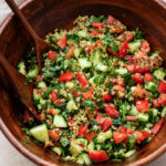 A wooden bowl filled with colorful tabbouleh salad made of chopped tomatoes, cucumbers, parsley, and bulgur, with wooden serving utensils.