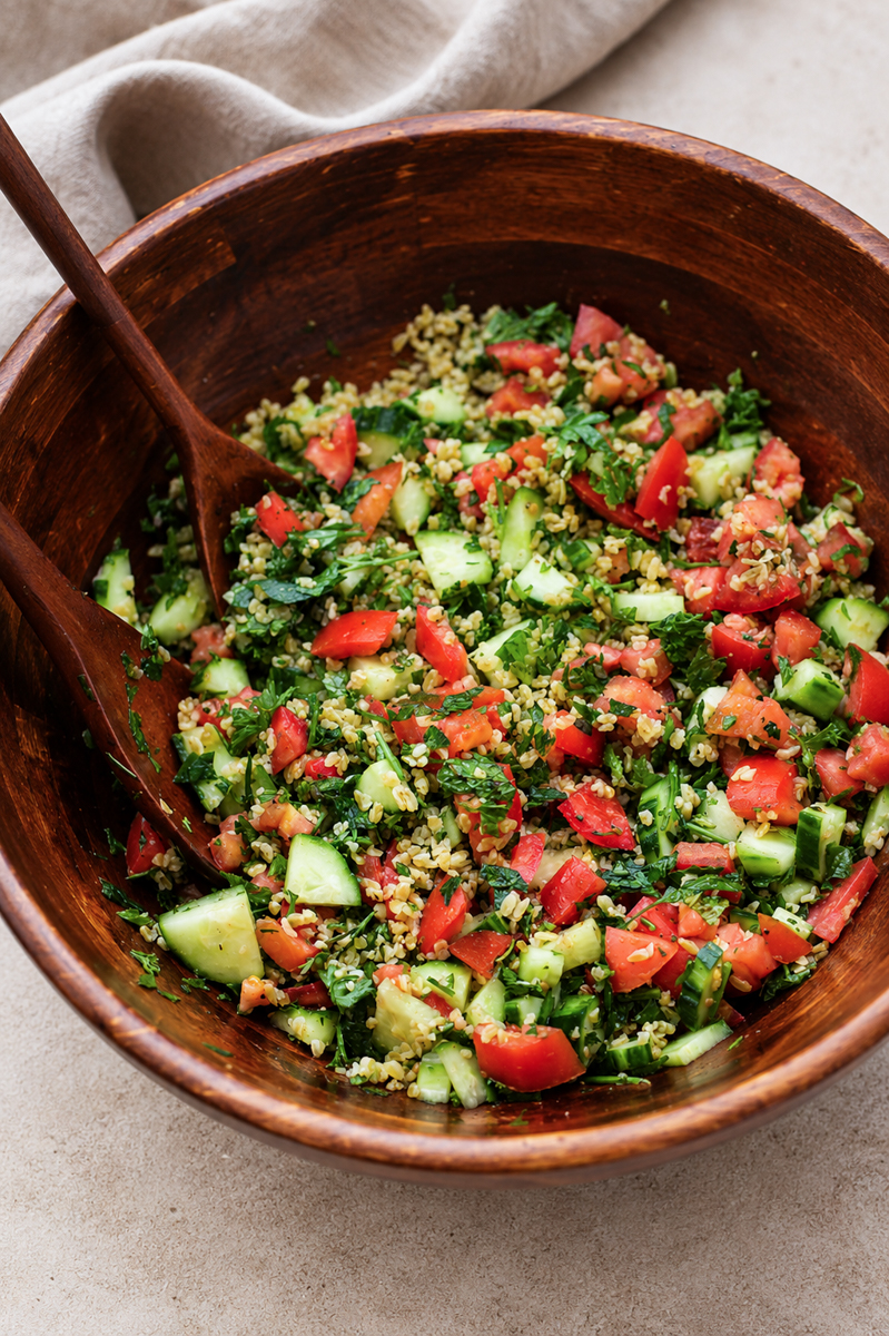 A wooden bowl filled with colorful tabbouleh salad made of chopped tomatoes, cucumbers, parsley, and bulgur, with wooden serving utensils.