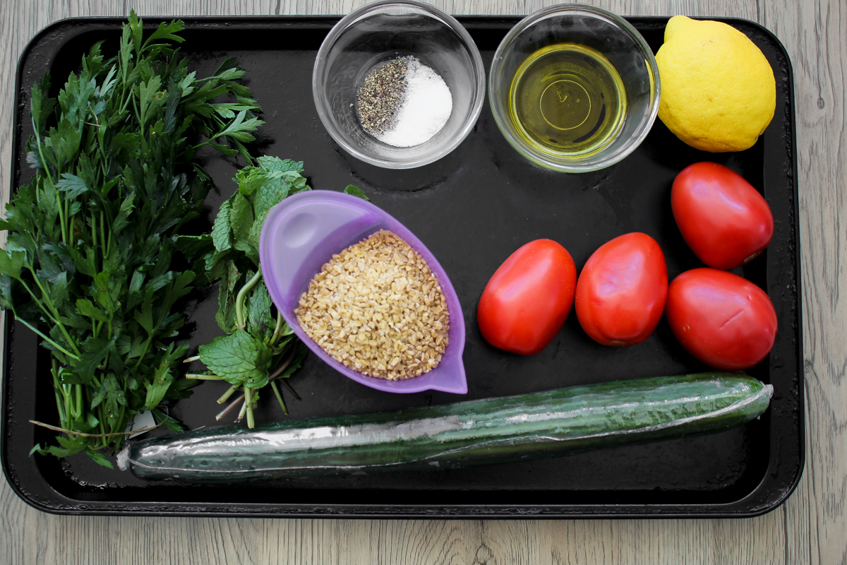 A tray with fresh parsley, mint, a cucumber, five Roma tomatoes, bulgur wheat in a purple bowl, a lemon, olive oil, and a small bowl of salt and pepper. All items are uncooked and neatly arranged.
