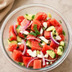 A glass bowl filled with a salad made of watermelon cubes, cucumber pieces, sliced red onion, crumbled feta cheese, and fresh mint on a light-colored surface.
