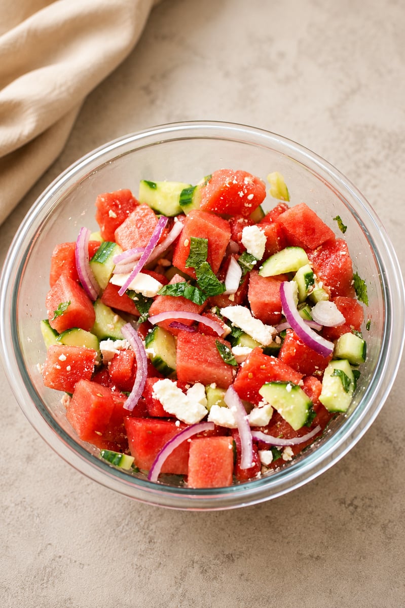 A glass bowl filled with a salad made of watermelon cubes, cucumber pieces, sliced red onion, crumbled feta cheese, and fresh mint on a light-colored surface.