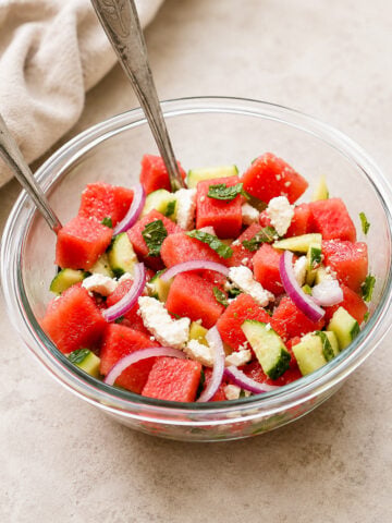 A glass bowl filled with a colorful salad of watermelon cubes, cucumber, red onion slices, crumbled feta cheese, and fresh herbs, with two metal spoons resting inside.