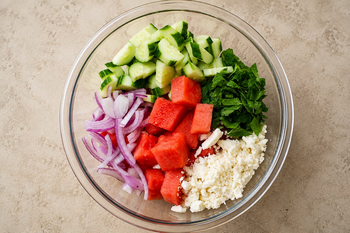 A glass bowl containing chopped cucumber, fresh mint, sliced red onion, cubed watermelon, and crumbled feta cheese arranged in separate sections on a beige surface.