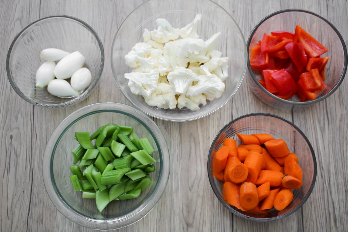 Five glass bowls with raw vegetables on a light wooden surface: peeled onions, cauliflower florets, red bell pepper pieces, celery slices, and carrot rounds.