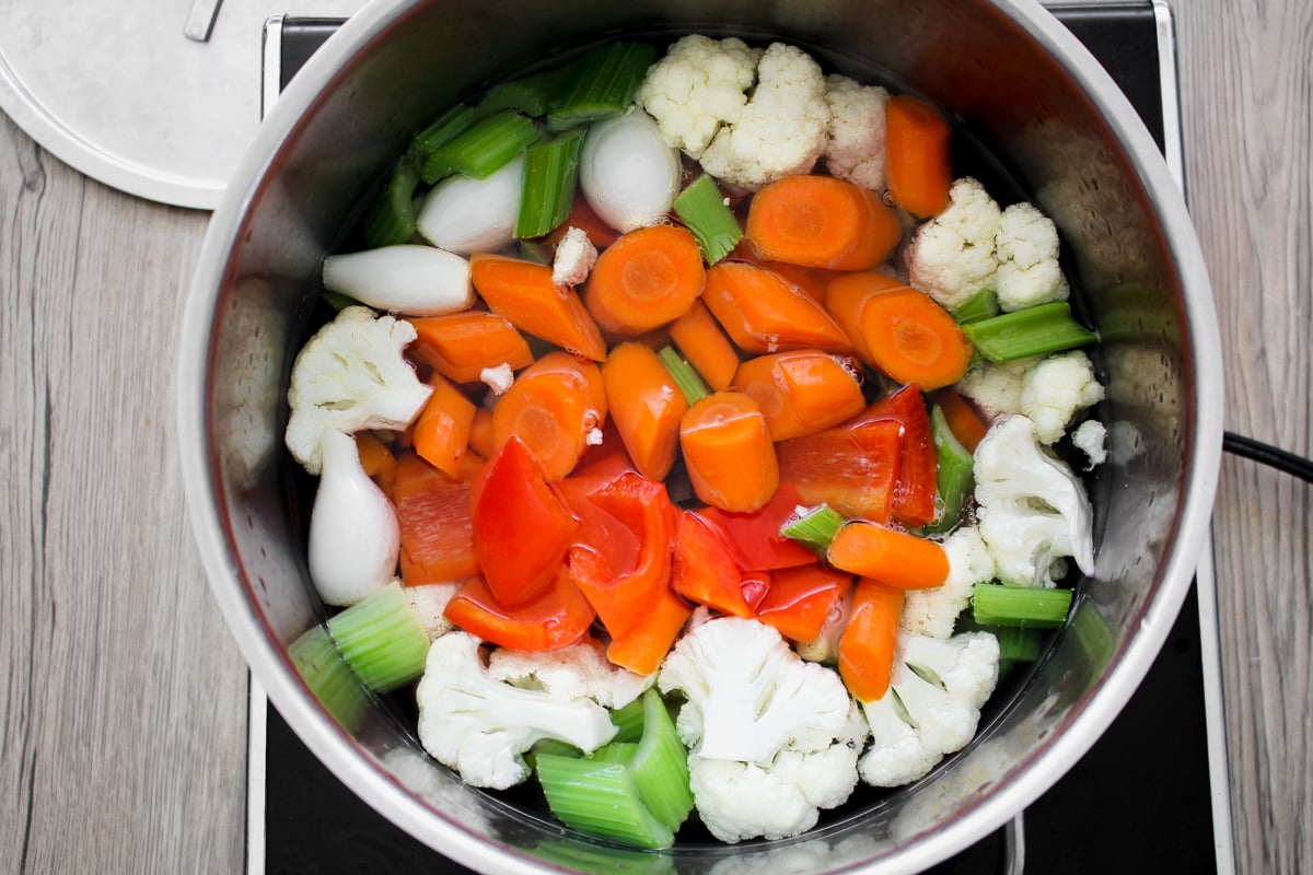 A pot filled with raw vegetables including carrots, celery, cauliflower, red bell pepper, and onions, sitting on a stovetop, ready to be cooked.