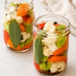Two glass jars filled with pickled vegetables, including cauliflower, carrots, celery, red peppers, bay leaves, and peppercorns, sit on a light surface with a beige cloth in the background.