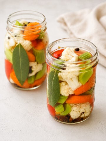 Two glass jars filled with pickled vegetables, including cauliflower, carrots, celery, red peppers, bay leaves, and peppercorns, sit on a light surface with a beige cloth in the background.