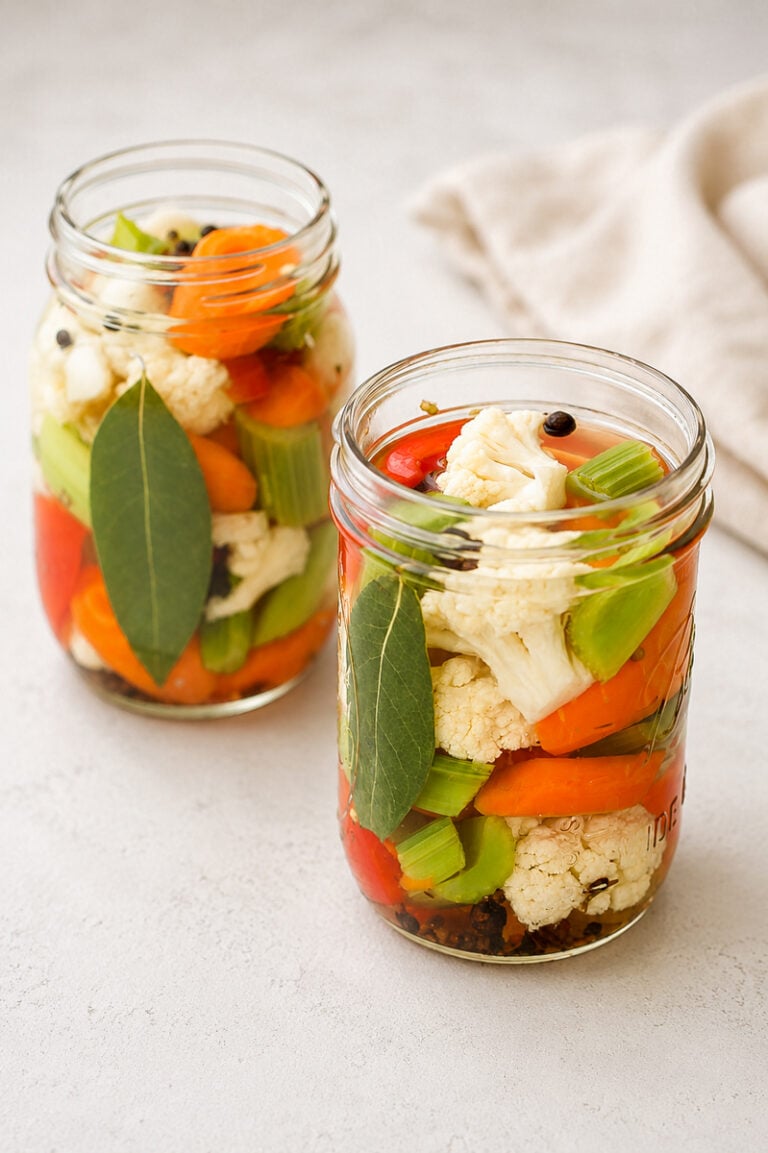 Two glass jars filled with pickled vegetables, including cauliflower, carrots, celery, red peppers, bay leaves, and peppercorns, sit on a light surface with a beige cloth in the background.