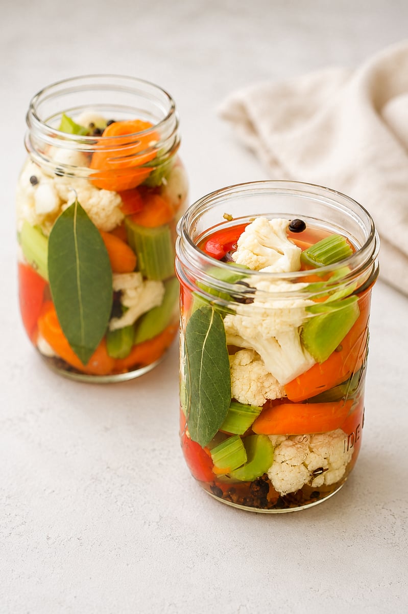 Two glass jars filled with pickled vegetables, including cauliflower, carrots, celery, red peppers, bay leaves, and peppercorns, sit on a light surface with a beige cloth in the background.