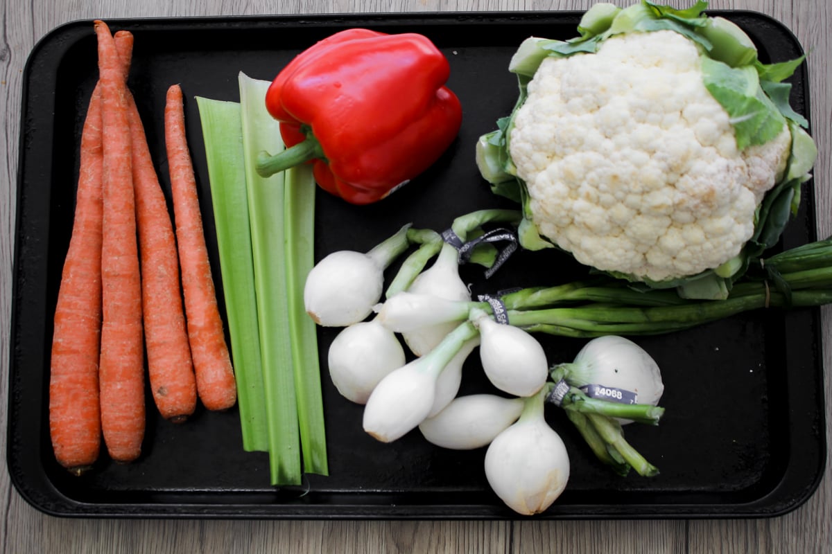 A tray with five carrots, two celery stalks, a red bell pepper, a head of cauliflower, and several white onions with green stems, all arranged neatly on a black tray.