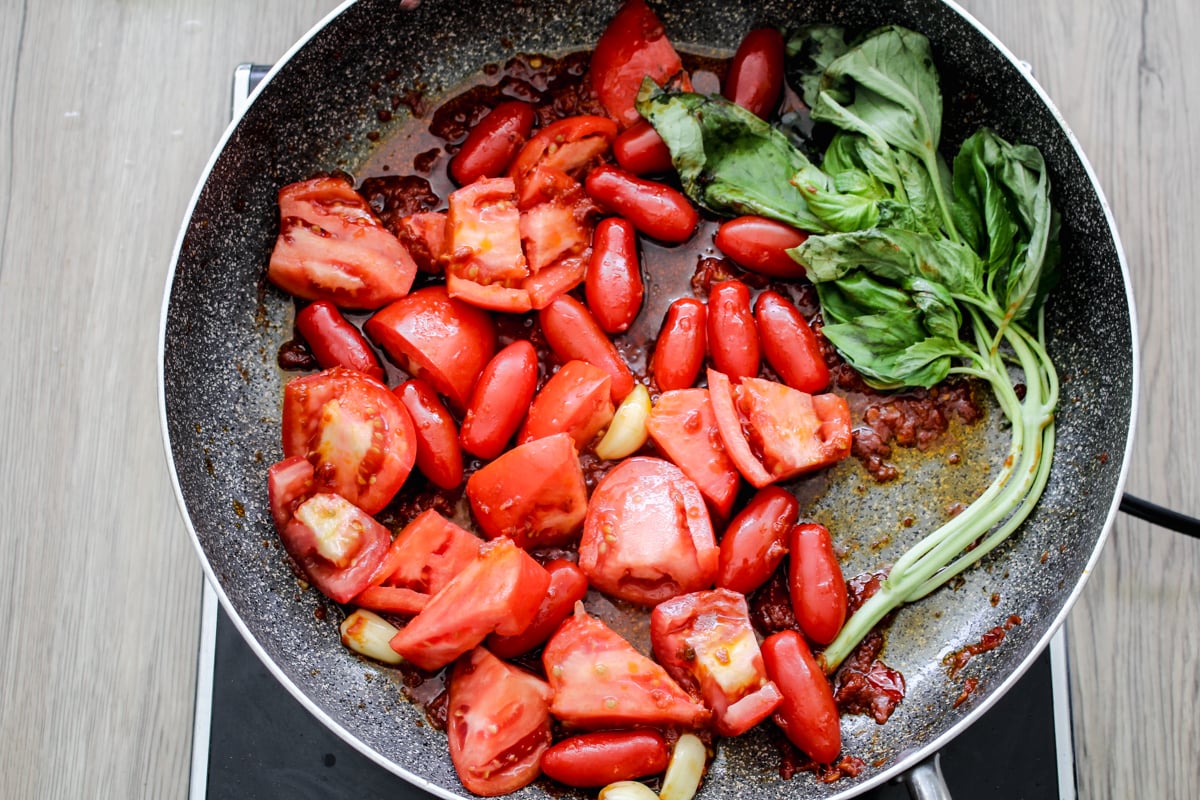 Tomatoes, garlic cloves, and fresh basil leaves are cooking in a frying pan on a stovetop, with visible tomato sauce and herbs.