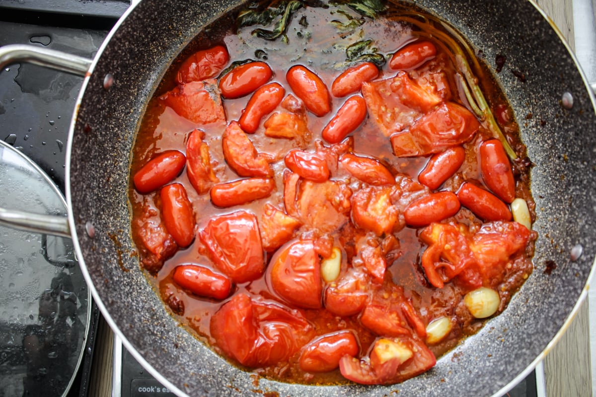 A pan filled with simmering tomatoes, garlic cloves, and herbs in a rich red sauce, cooking on a stovetop.