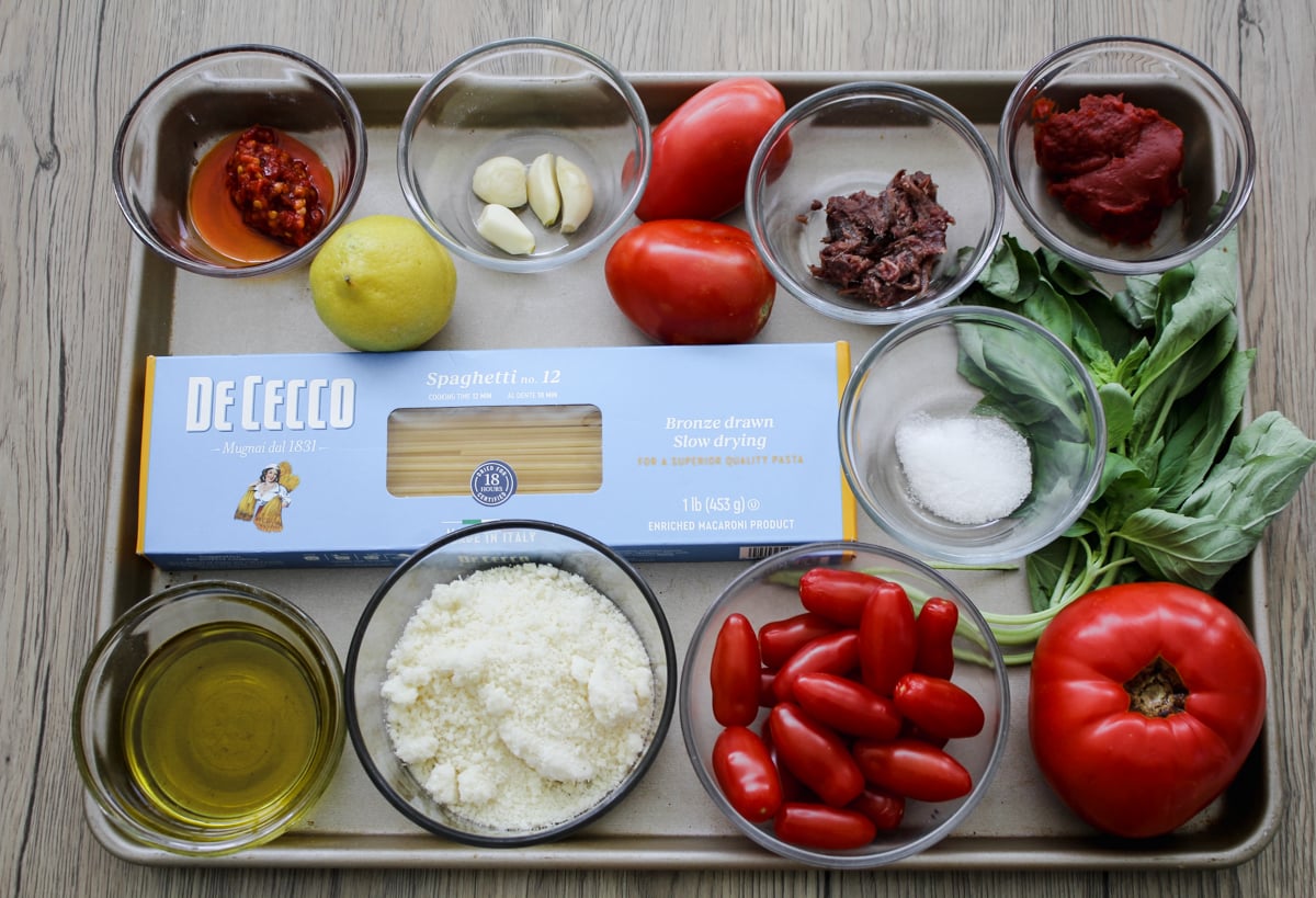 A tray holds spaghetti, cherry and large tomatoes, basil, olive oil, grated cheese, lemon, garlic, red pepper, chopped olives, tomato paste, salt, and chili paste in small bowls on a wooden surface.