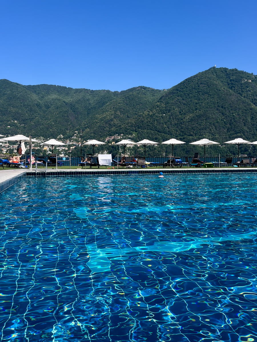 A clear blue swimming pool with sun loungers and white umbrellas along the edge, mountains and green hills in the background under a bright blue sky.