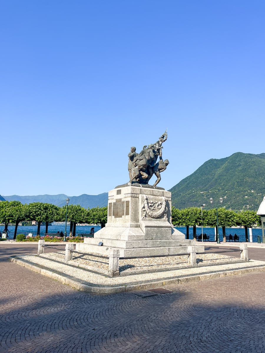 A bronze equestrian statue on a stone pedestal stands in a plaza near a lake, with green mountains and blue sky in the background.