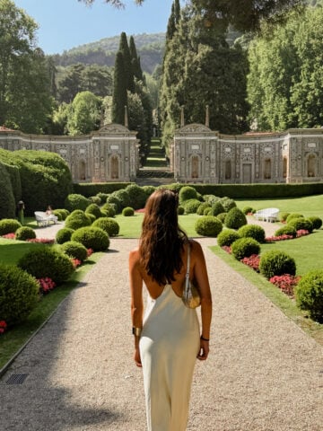 A woman in a backless dress stands on a gravel path in a manicured garden, facing ornate stone buildings, trees, and greenery under a sunny sky.
