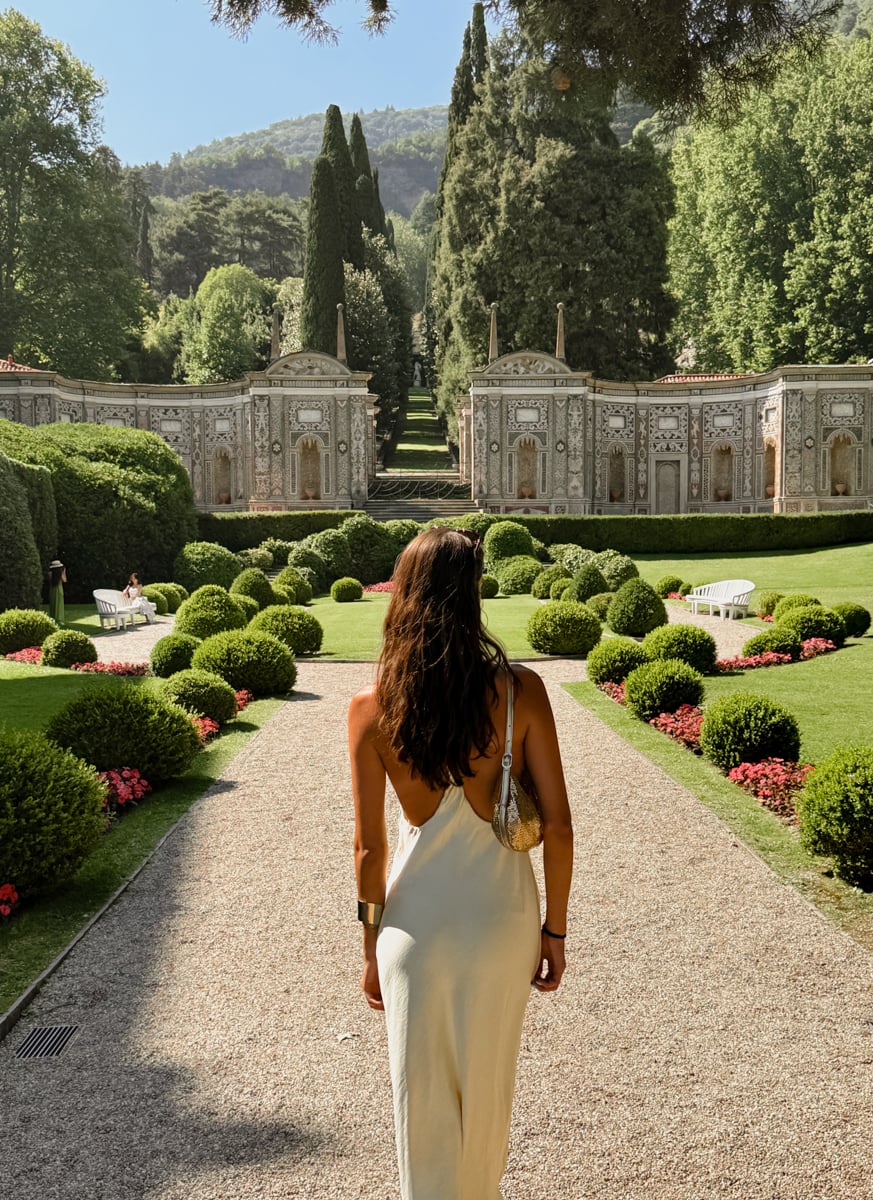 A woman in a backless dress stands on a gravel path in a manicured garden, facing ornate stone buildings, trees, and greenery under a sunny sky.
