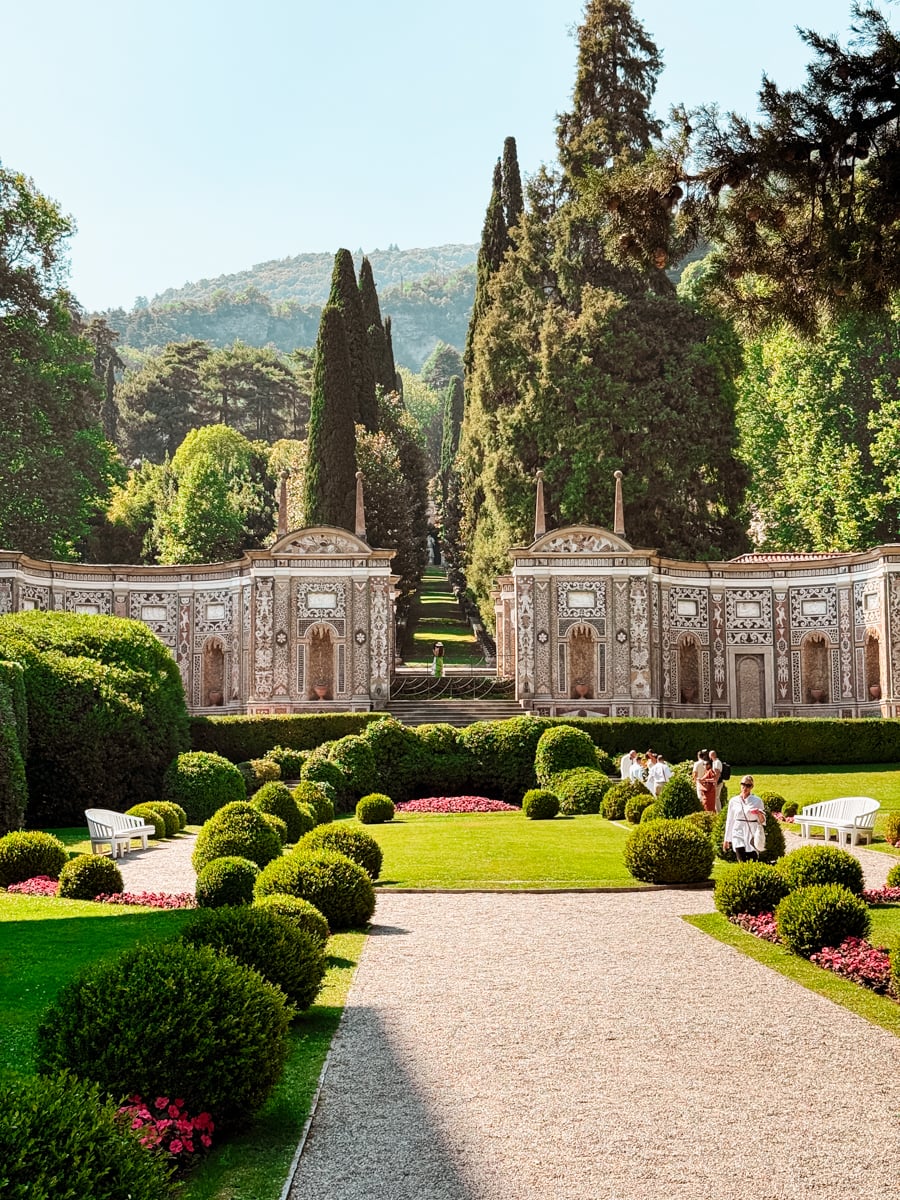 Ornate garden with manicured bushes, gravel path, benches, and groups of people. Decorative stone wall and tall trees frame a scenic backdrop of hills under a clear sky.
