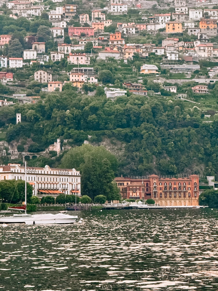Colorful hillside buildings overlook a lake with boats docked along the shore. Lush green trees cover the slope between the water and houses.