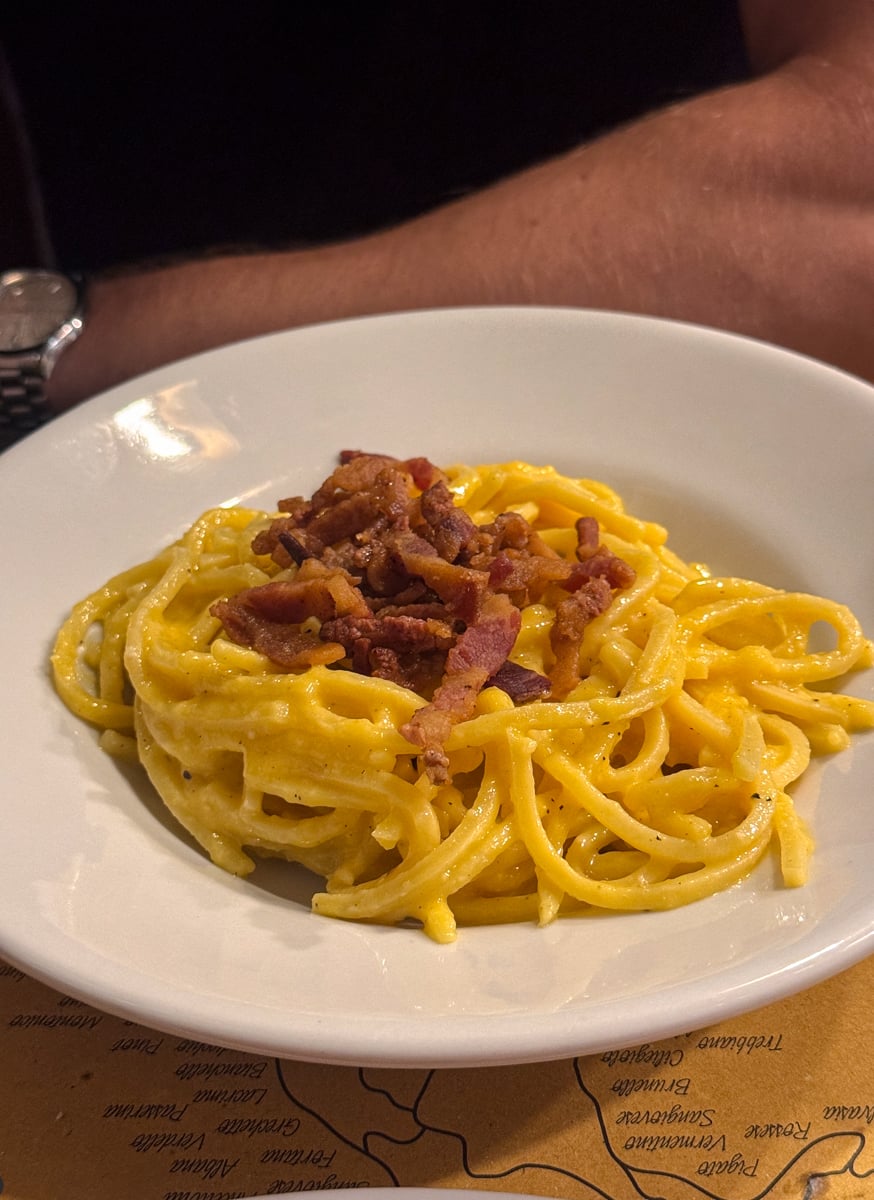 A white plate of spaghetti carbonara topped with crispy bacon pieces is in the foreground, with a persons arm and a partial map visible in the background.