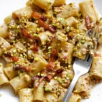 A white plate with rigatoni pasta in a creamy sauce, topped with bacon pieces and chopped pistachios. A metal fork rests on the side of the plate.