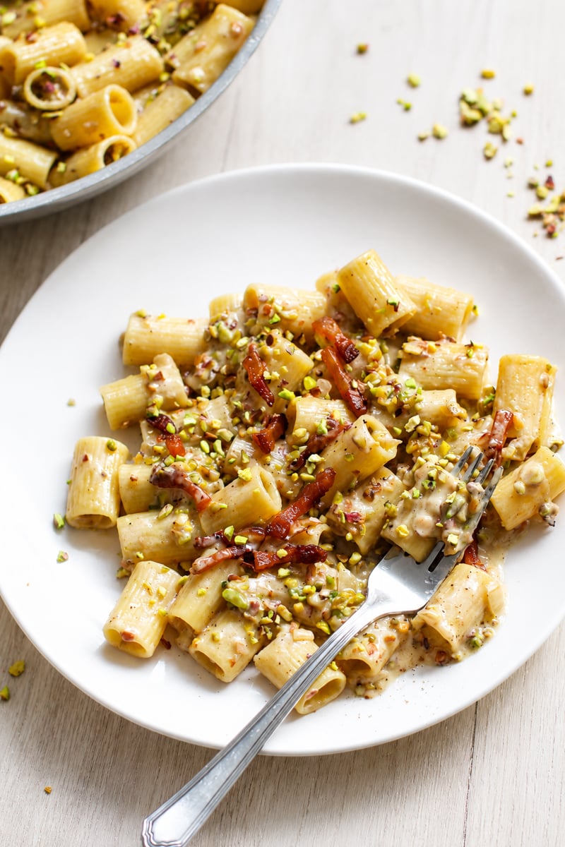 Plate of rigatoni pasta topped with creamy sauce, pieces of crispy bacon, and chopped pistachios, with a fork resting on the plate. A pan with more pasta is partially visible in the background.