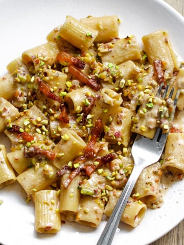 A white plate with rigatoni pasta in a creamy sauce, topped with bacon pieces and chopped pistachios. A metal fork rests on the side of the plate.