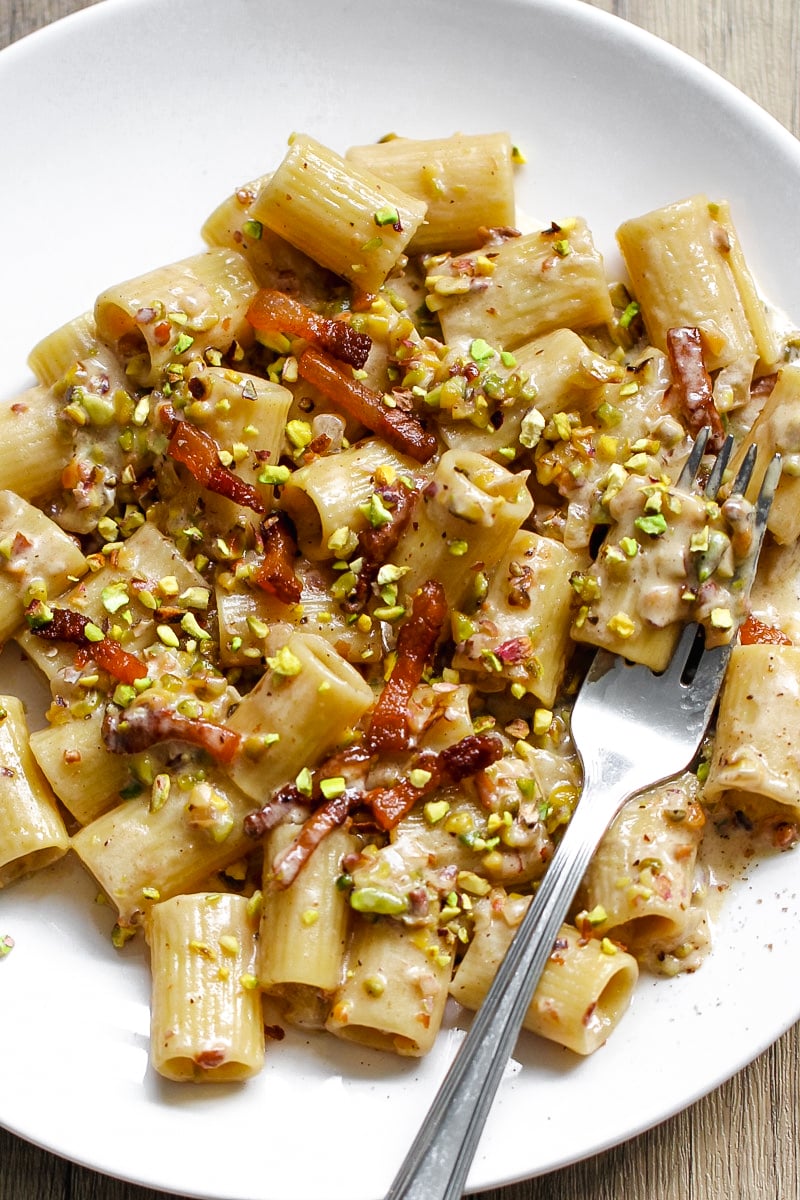 A white plate with rigatoni pasta in a creamy sauce, topped with bacon pieces and chopped pistachios. A metal fork rests on the side of the plate.