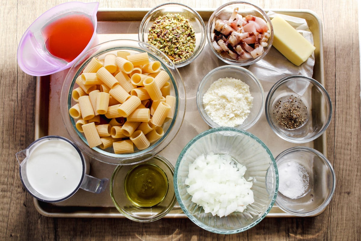 A tray with bowls containing rigatoni pasta, chopped onions, diced bacon, grated cheese, pistachios, butter, cream, olive oil, pink sauce, salt, and pepper on a wooden surface.