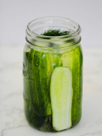 A clear glass jar filled with whole and sliced pickles, sitting on a white surface with a light background.