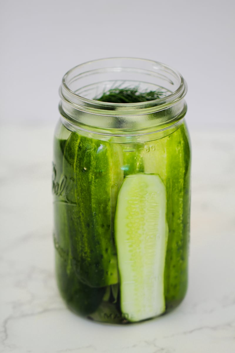 A clear glass jar filled with whole and sliced pickles, sitting on a white surface with a light background.