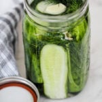 A glass jar filled with cucumber spears, dill, and garlic cloves in brine sits on a marble surface next to a striped towel and a metal jar lid.