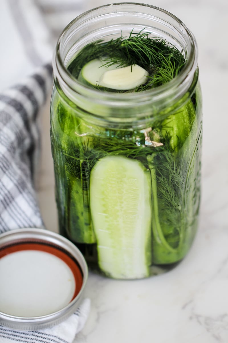 A glass jar filled with cucumber spears, dill, and garlic cloves in brine sits on a marble surface next to a striped towel and a metal jar lid.